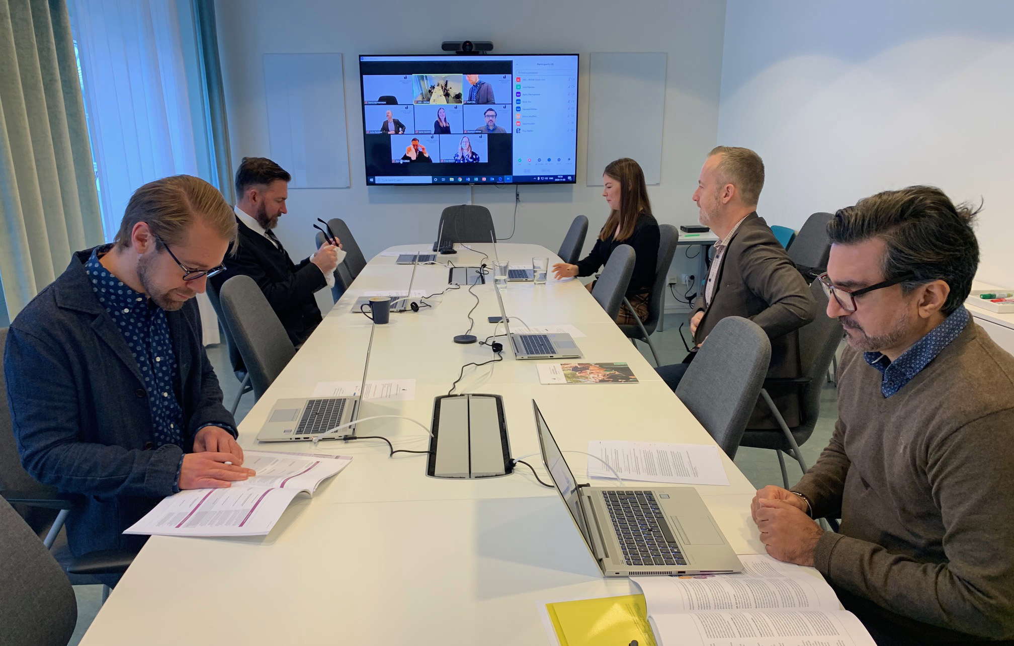 JIBS staff in meeting room with laptops. Projector screen in background with zoom meeting participants showing.