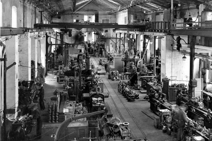A historic photo of the inside of a factory hall in black & white. There are lots of different machines throughout the factory floor except in the middle where the floor is cleared to make room for a transport rail. In the roof a large crane is mounted.