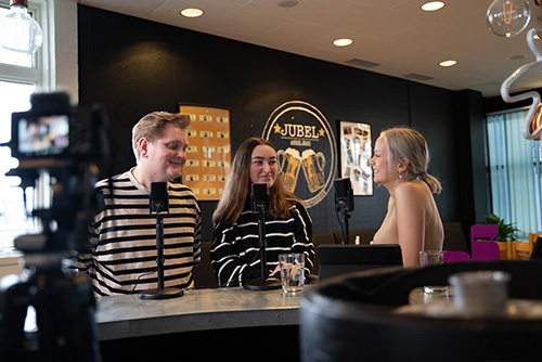 Three students sit in conversation in a campus setting. They have microphones in front of them and cameras in the foreground filming them.